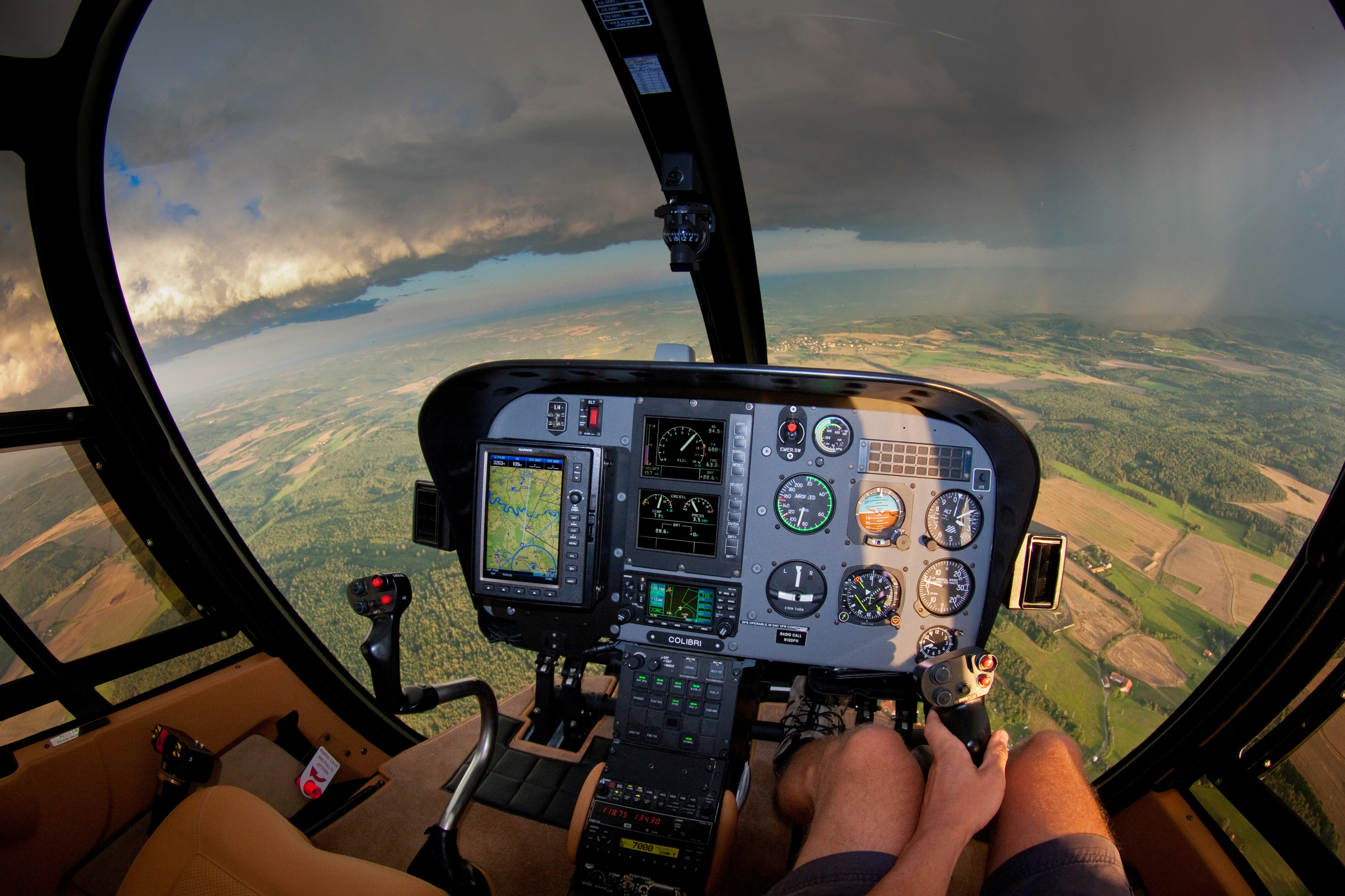 Helicopter cockpit with avionics glowing, flying toward dramatic storm clouds over green terrain